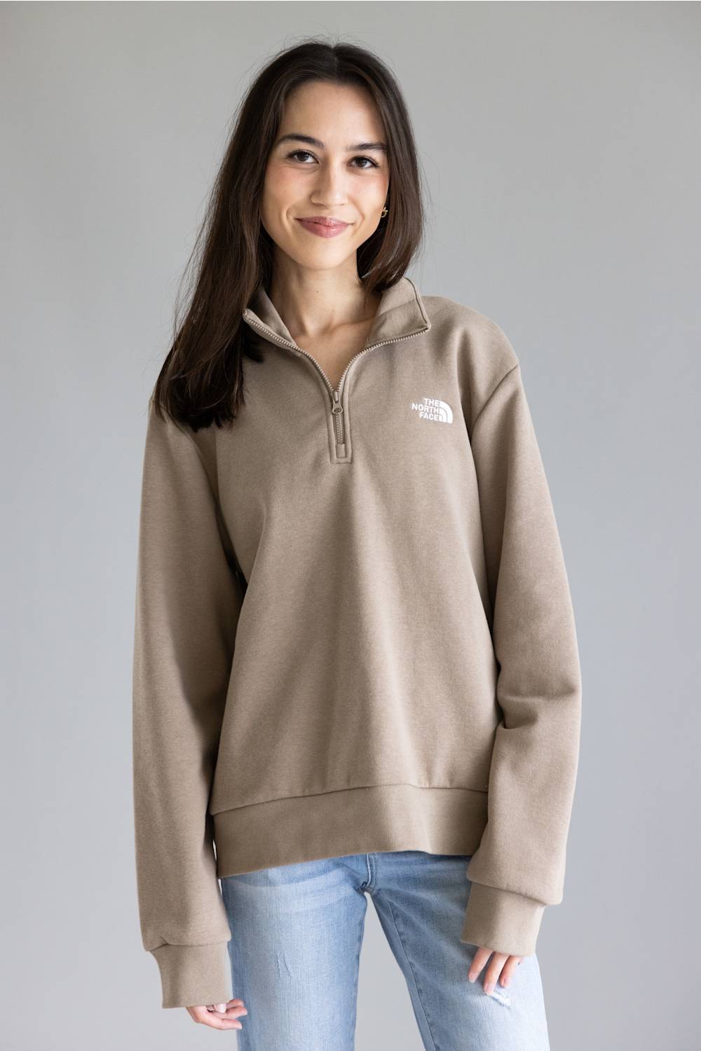 A woman wearing a taupe brown quarter-zip pullover sweatshirt by The North Face, paired with light blue jeans, standing against a neutral gray studio background.
