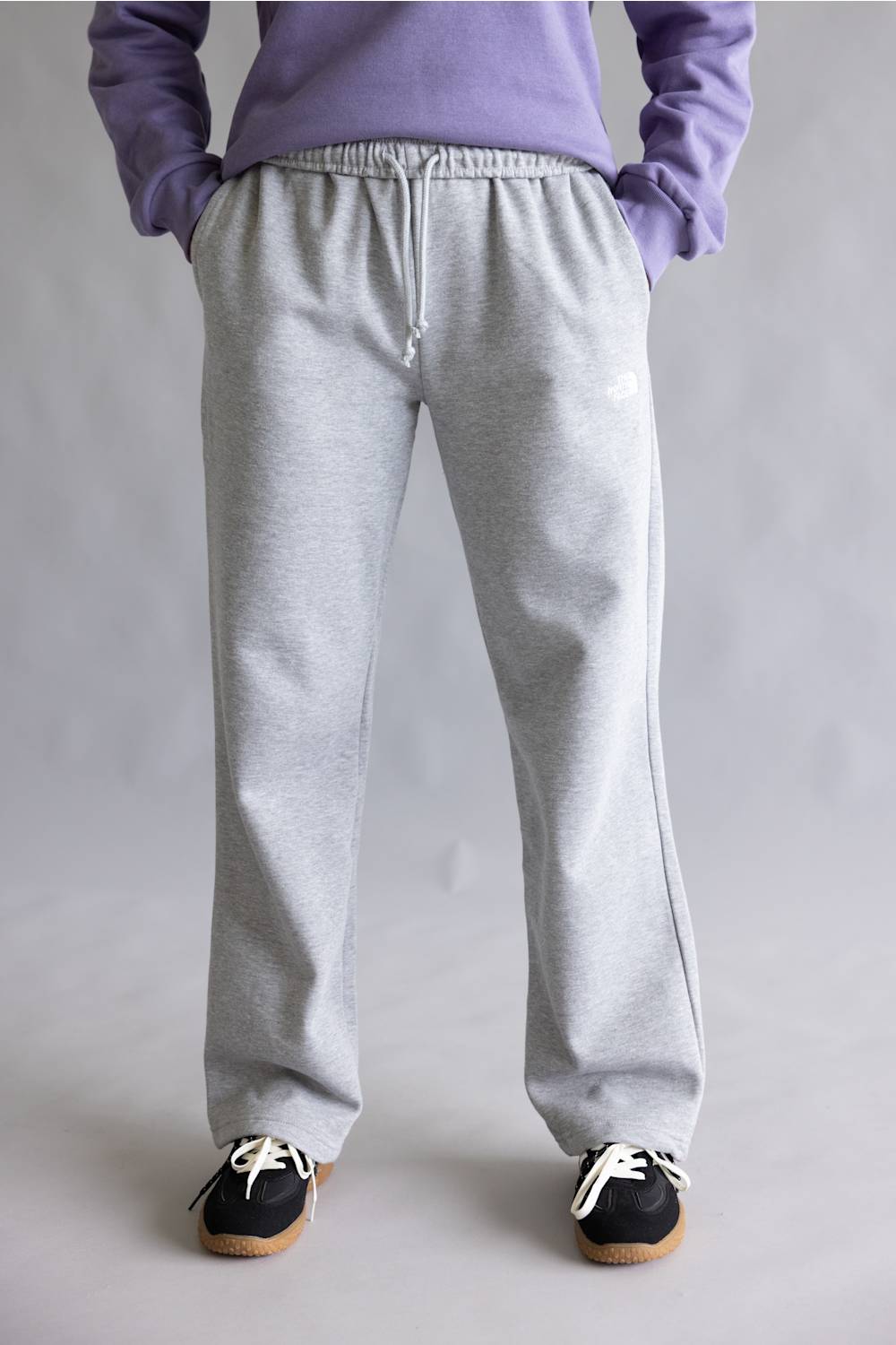 A person shown from the waist down wearing light gray heather sweatpants, a lavender sweatshirt, and black sneakers against a neutral gray studio background.