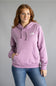 A woman wearing a light purple/lavender pullover hoodie with a small white embroidered logo on the left chest, paired with blue jeans, standing against a solid gray studio background.