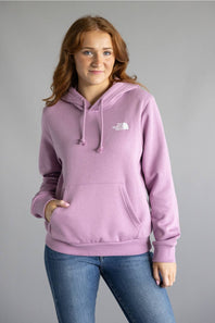 A woman wearing a light purple/lavender pullover hoodie with a small white embroidered logo on the left chest, paired with blue jeans, standing against a solid gray studio background.