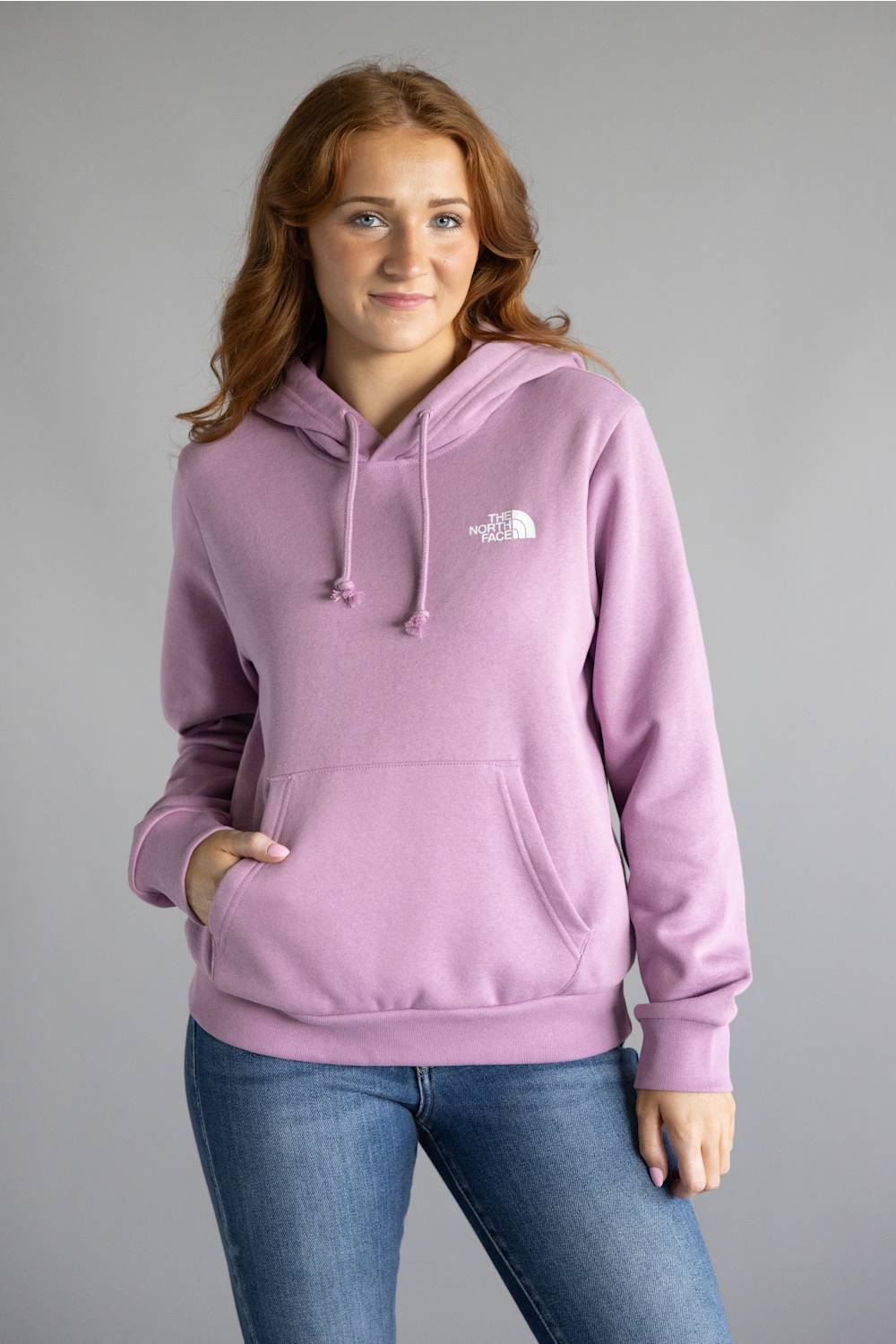 A woman wearing a light purple/lavender pullover hoodie with a small white embroidered logo on the left chest, paired with blue jeans, standing against a solid gray studio background.