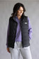 A woman wearing a black puffer vest by The North Face over a purple quarter-zip pullover and light gray sweatpants, standing against a neutral gray studio background.