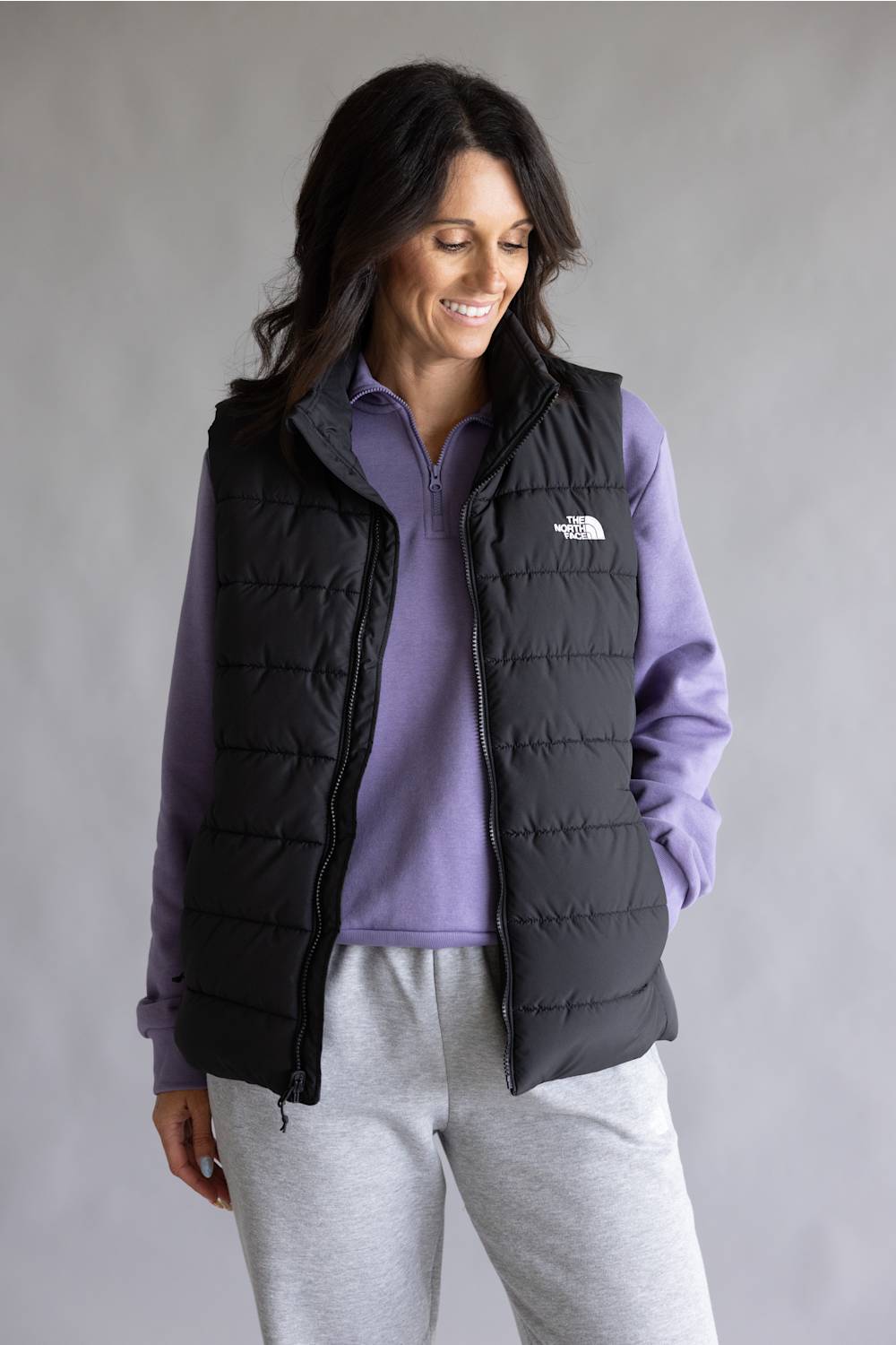 A woman wearing a black puffer vest by The North Face over a purple quarter-zip pullover and light gray sweatpants, standing against a neutral gray studio background.