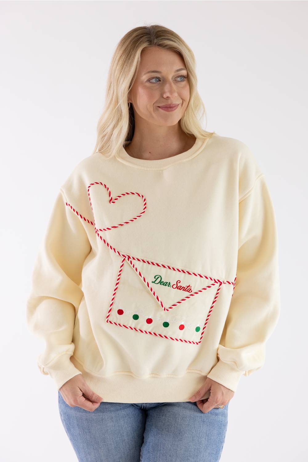 A woman wearing a cream-colored crewneck sweatshirt with a red and green Christmas-themed graphic of an envelope, a heart, and the text 'Dear Santa'.