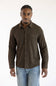 A man wearing a dark brown long-sleeve button-up shirt with a chest pocket, paired with light blue jeans, standing against a plain white background.