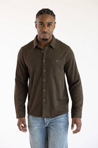 A man wearing a dark brown long-sleeve button-up shirt with a chest pocket, paired with light blue jeans, standing against a plain white background.