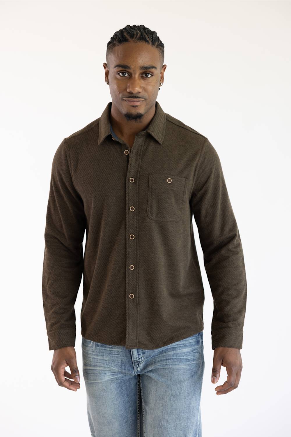 A man wearing a dark brown long-sleeve button-up shirt with a chest pocket, paired with light blue jeans, standing against a plain white background.