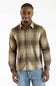A man wearing a brown and beige plaid long-sleeve button-down shirt with blue jeans, standing against a plain white background.
