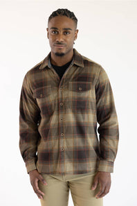 A man wearing a brown and beige plaid button-down shirt over a black undershirt, paired with khaki pants, standing against a plain white background.
