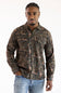 A man wearing a long-sleeve button-up shirt with an all-over camouflage pattern in shades of brown, green, and black, paired with blue jeans, against a plain white background.