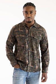 A man wearing a long-sleeve button-up shirt with an all-over camouflage pattern in shades of brown, green, and black, paired with blue jeans, against a plain white background.