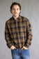 A man wearing a brown and black plaid button-down flannel shirt with blue jeans, standing against a neutral gray studio background.