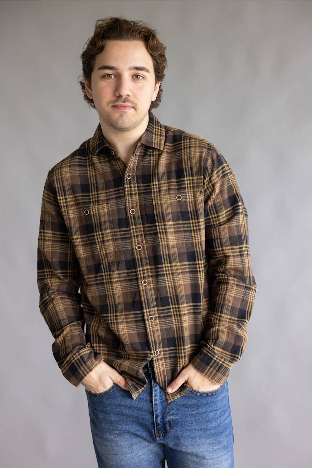 A man wearing a brown and black plaid button-down flannel shirt with blue jeans, standing against a neutral gray studio background.