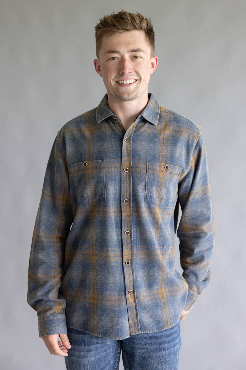 A smiling man wearing a long-sleeve button-up plaid flannel shirt in slate blue and mustard yellow, paired with blue jeans, standing against a neutral gray studio background.