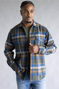 A man wearing a long-sleeve plaid button-up shirt in blue, green, and beige over a dark olive t-shirt and blue jeans, standing against a neutral gray studio background.