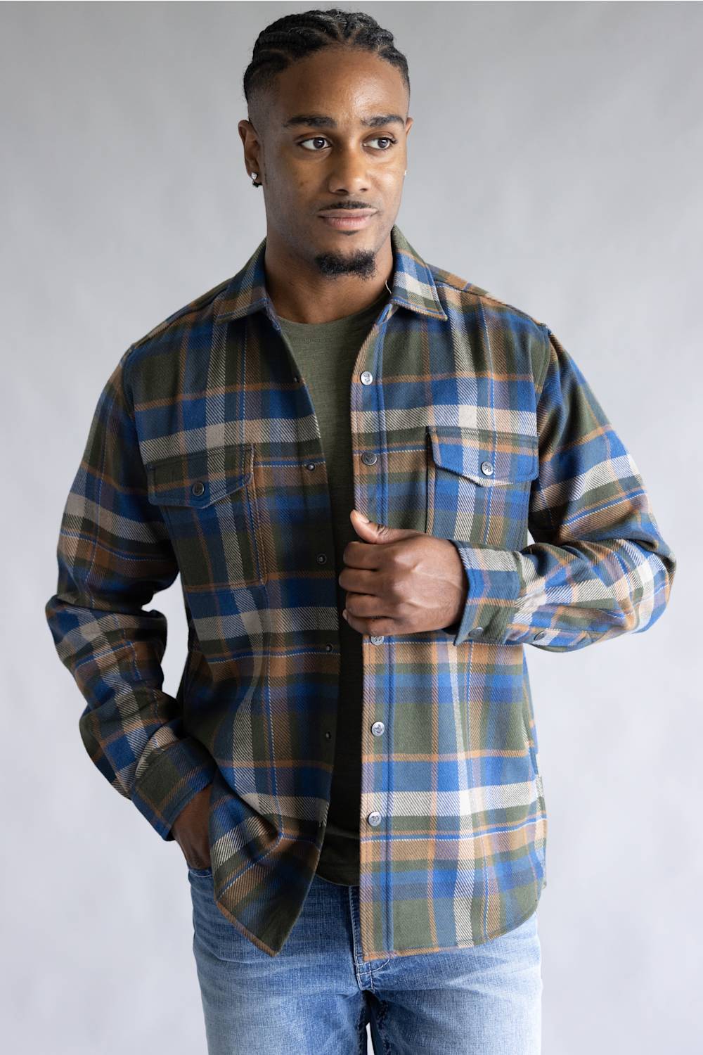 A man wearing a long-sleeve plaid button-up shirt in blue, green, and beige over a dark olive t-shirt and blue jeans, standing against a neutral gray studio background.