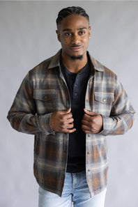 A man wearing a brown and beige plaid flannel shirt over a black henley t-shirt and light blue jeans, standing against a neutral gray studio background.