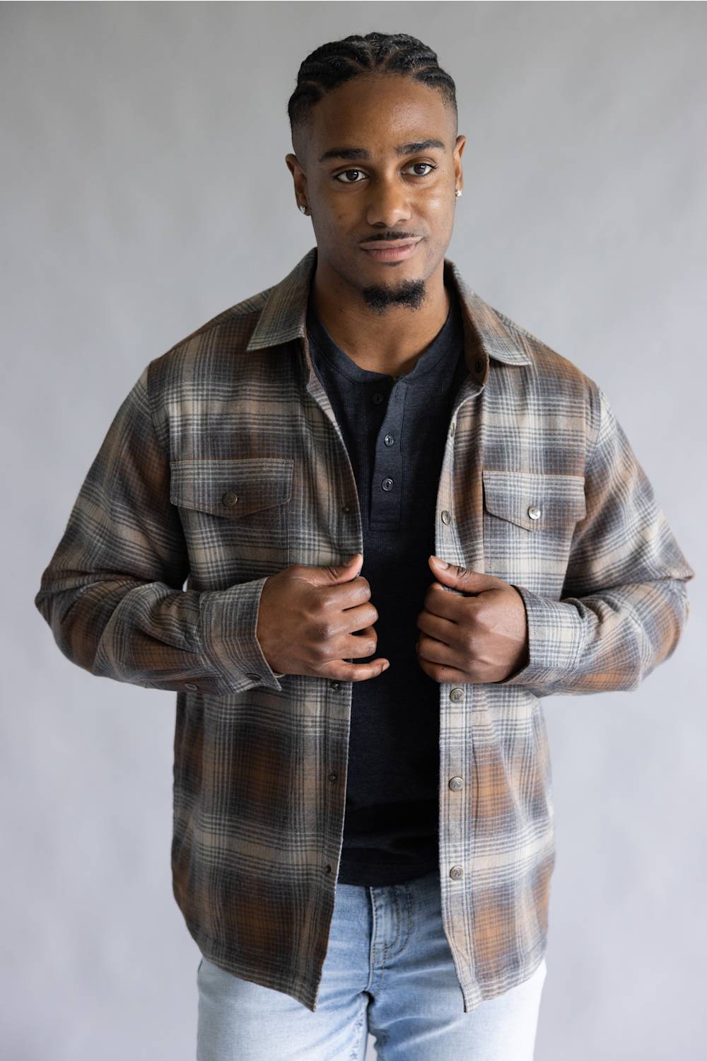 A man wearing a brown and beige plaid flannel shirt over a black henley t-shirt and light blue jeans, standing against a neutral gray studio background.