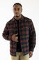 A man wearing a dark plaid button-up shirt jacket over a black t-shirt and khaki pants, standing against a plain white background.