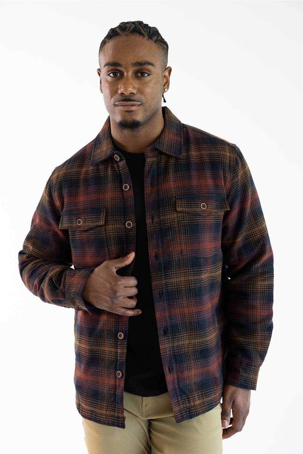 A man wearing a dark plaid button-up shirt jacket over a black t-shirt and khaki pants, standing against a plain white background.