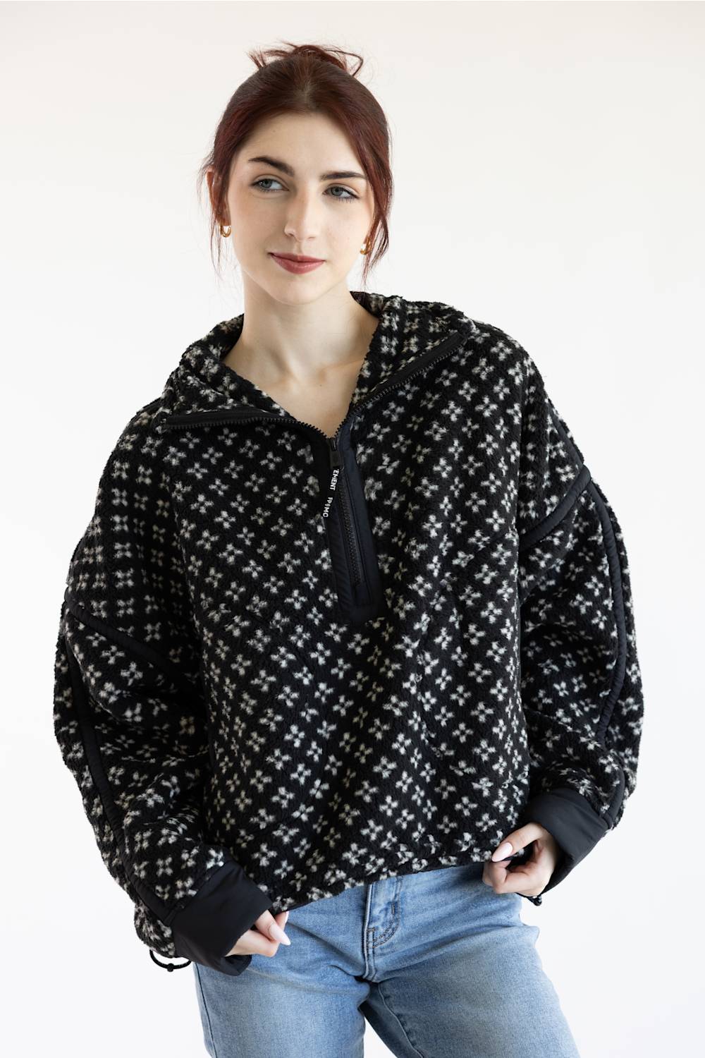 A woman wearing a black and white patterned half-zip hoodie with a hood and ribbed cuffs, paired with light blue jeans, photographed against a plain white studio background.
