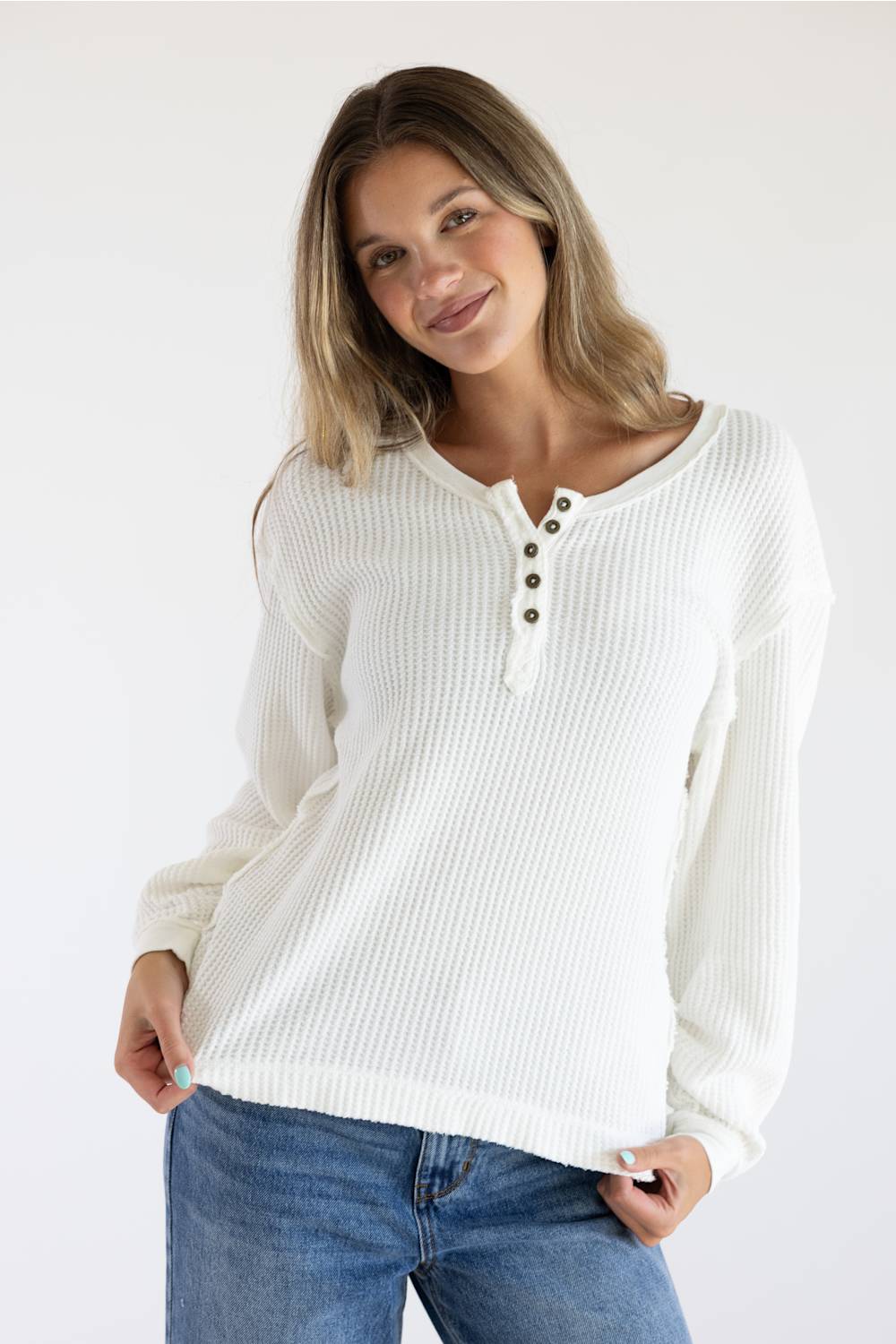A woman wearing a white ribbed long-sleeve henley top with three buttons, paired with blue jeans, against a plain white background.