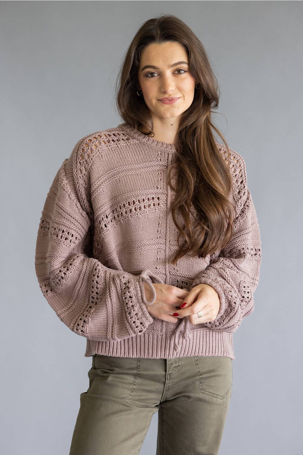 A woman modeling a dusty rose knit sweater with textured openwork patterns and ribbed cuffs, paired with olive green pants, against a solid gray studio background.
