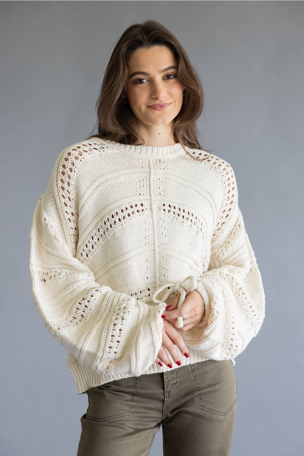 A woman wearing a cream-colored, textured knit sweater with intricate openwork patterns and voluminous sleeves, paired with olive green pants, set against a plain gray studio background.