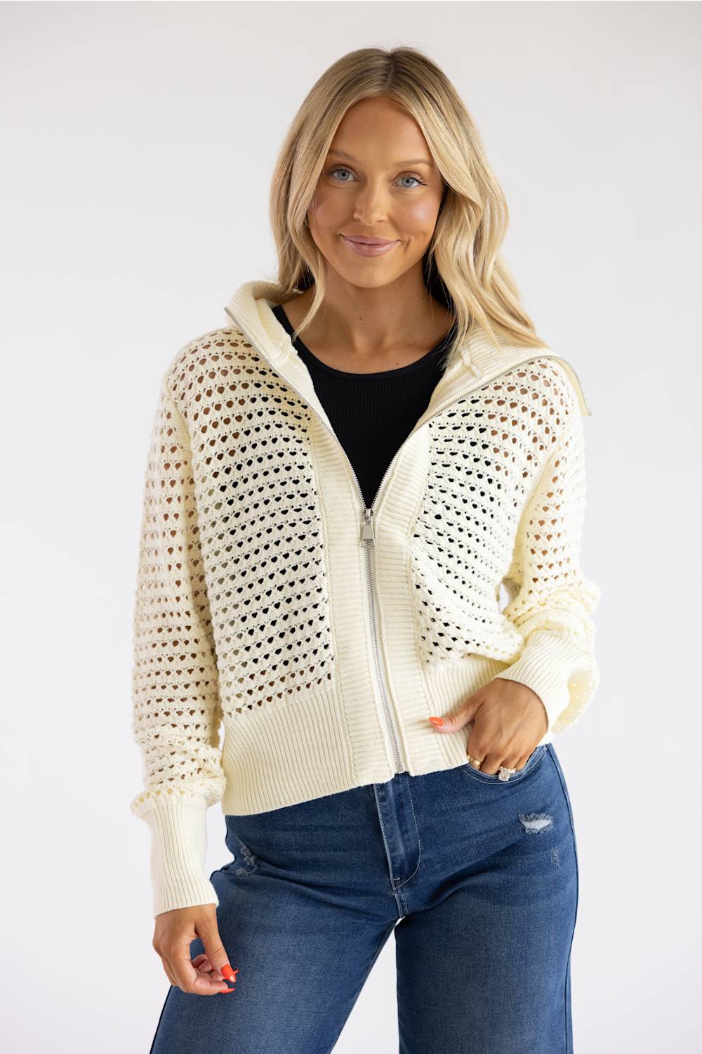 A woman modeling a cream open-knit zip-up hoodie jacket over a black top and blue jeans, photographed against a plain light gray studio background.