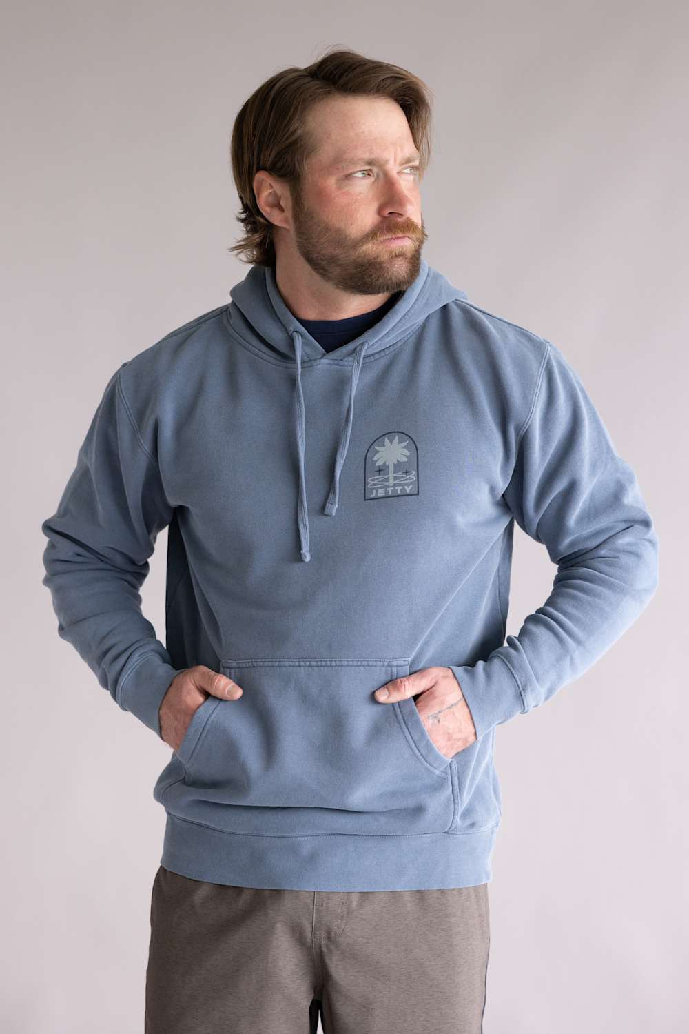 Man wearing a blue hoodie with a logo on a plain background. The hoodie is Jetty Orbit Grove Hoodie for Men in Pigment Slate