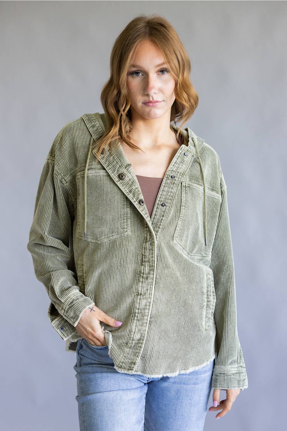 A woman wearing an oversized sage green corduroy button-up shirt jacket over a brown top and light blue jeans, standing against a neutral gray studio background.