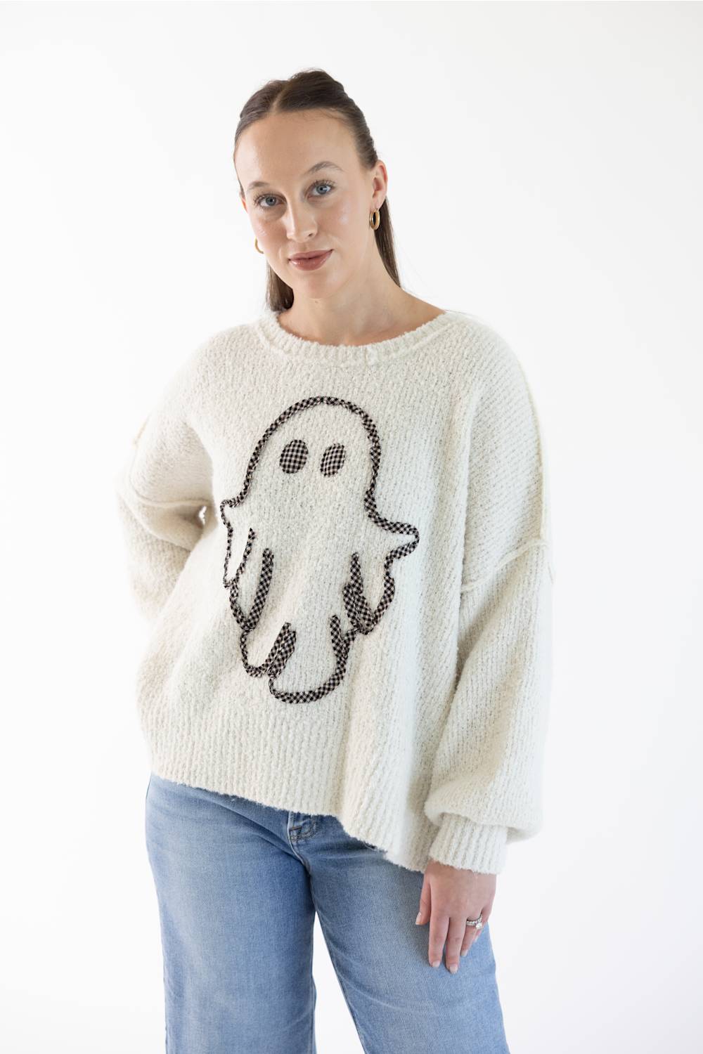 A woman wearing a cream-colored knit sweater with a black and light orange gingham ghost design on the front, paired with light blue jeans, against a plain white background.