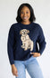 A smiling woman wearing a navy blue crewneck sweater with a large beige and cream dog graphic on the front, paired with light blue jeans, against a plain white studio background.