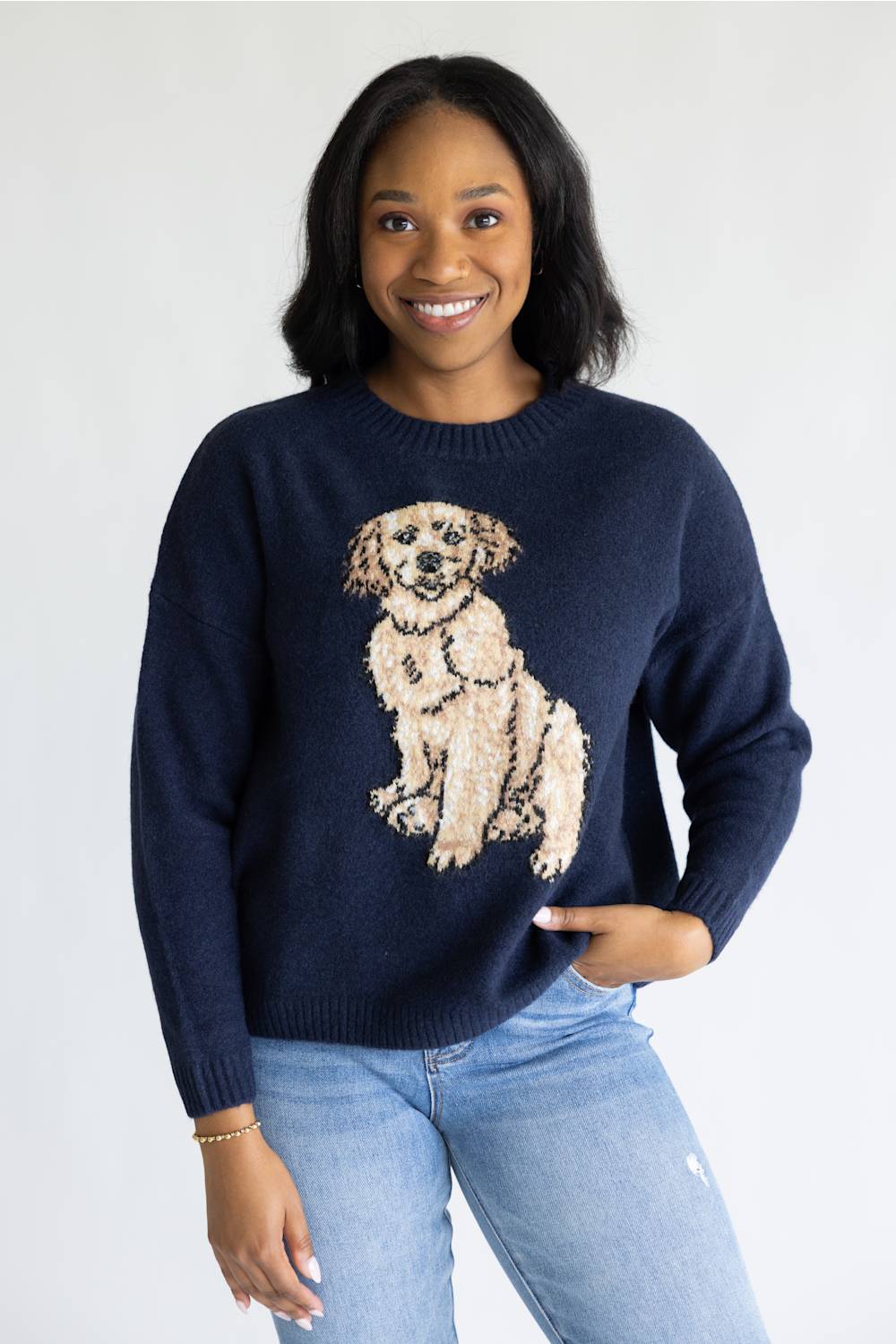 A smiling woman wearing a navy blue crewneck sweater with a large beige and cream dog graphic on the front, paired with light blue jeans, against a plain white studio background.