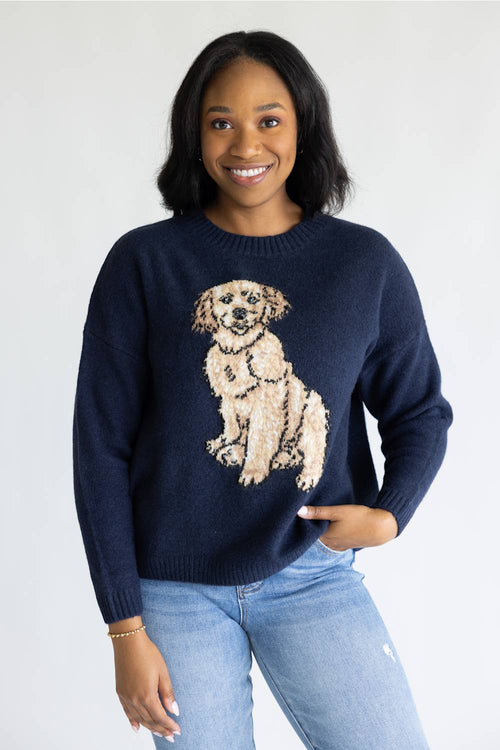A smiling woman wearing a navy blue crewneck sweater with a large beige and cream dog graphic on the front, paired with light blue jeans, against a plain white studio background.