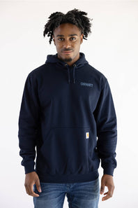 A man wearing a navy blue Carhartt pullover hoodie with the brand name printed on the left chest and a signature yellow logo patch on the pocket, paired with blue jeans against a plain white background.
