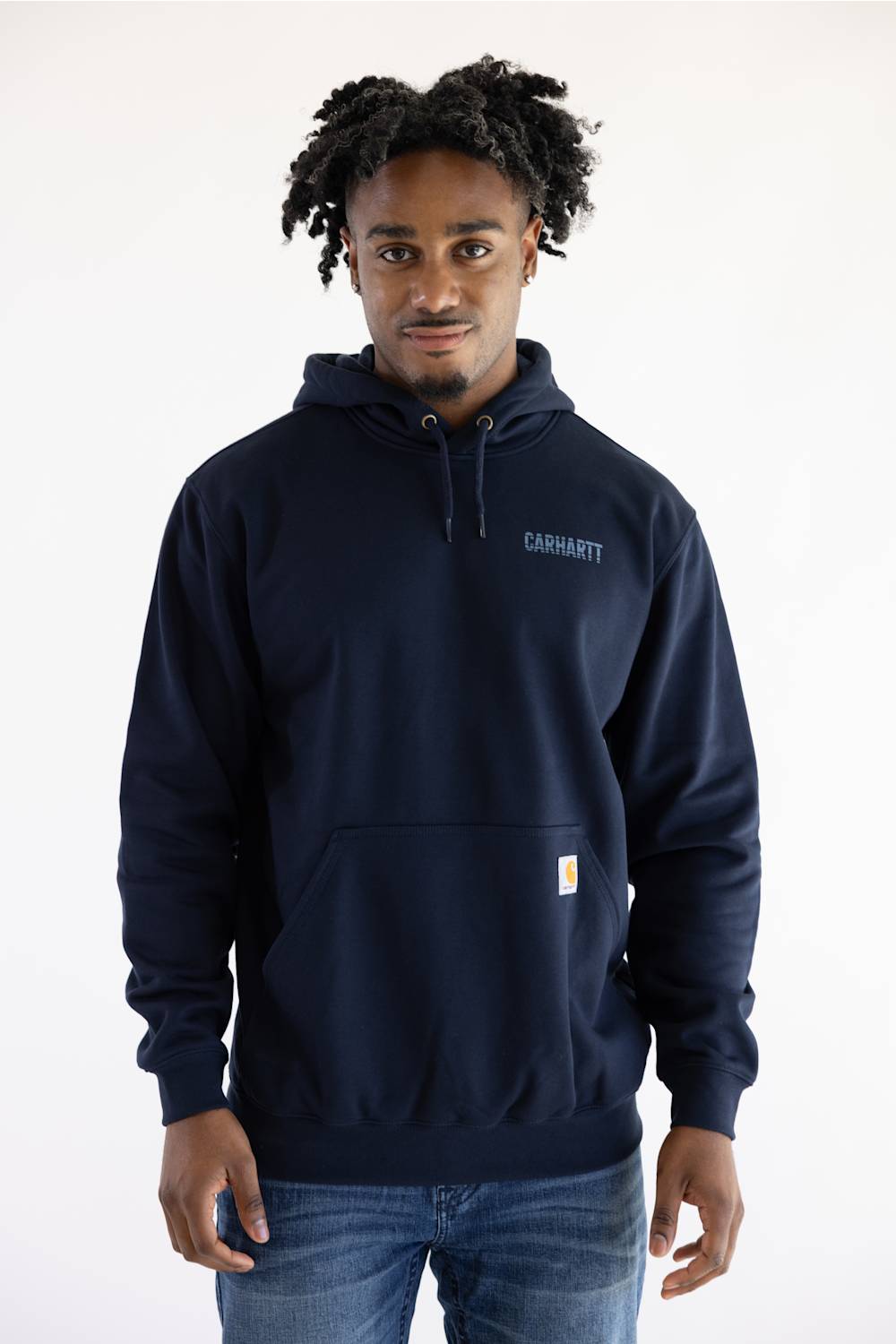 A man wearing a navy blue Carhartt pullover hoodie with the brand name printed on the left chest and a signature yellow logo patch on the pocket, paired with blue jeans against a plain white background.