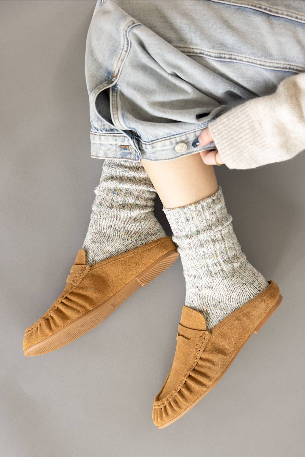 Side-angle lifestyle view of a person wearing Project Cloud tan suede backless loafer mules styled with thick cozy marled socks and cuffed light blue denim.