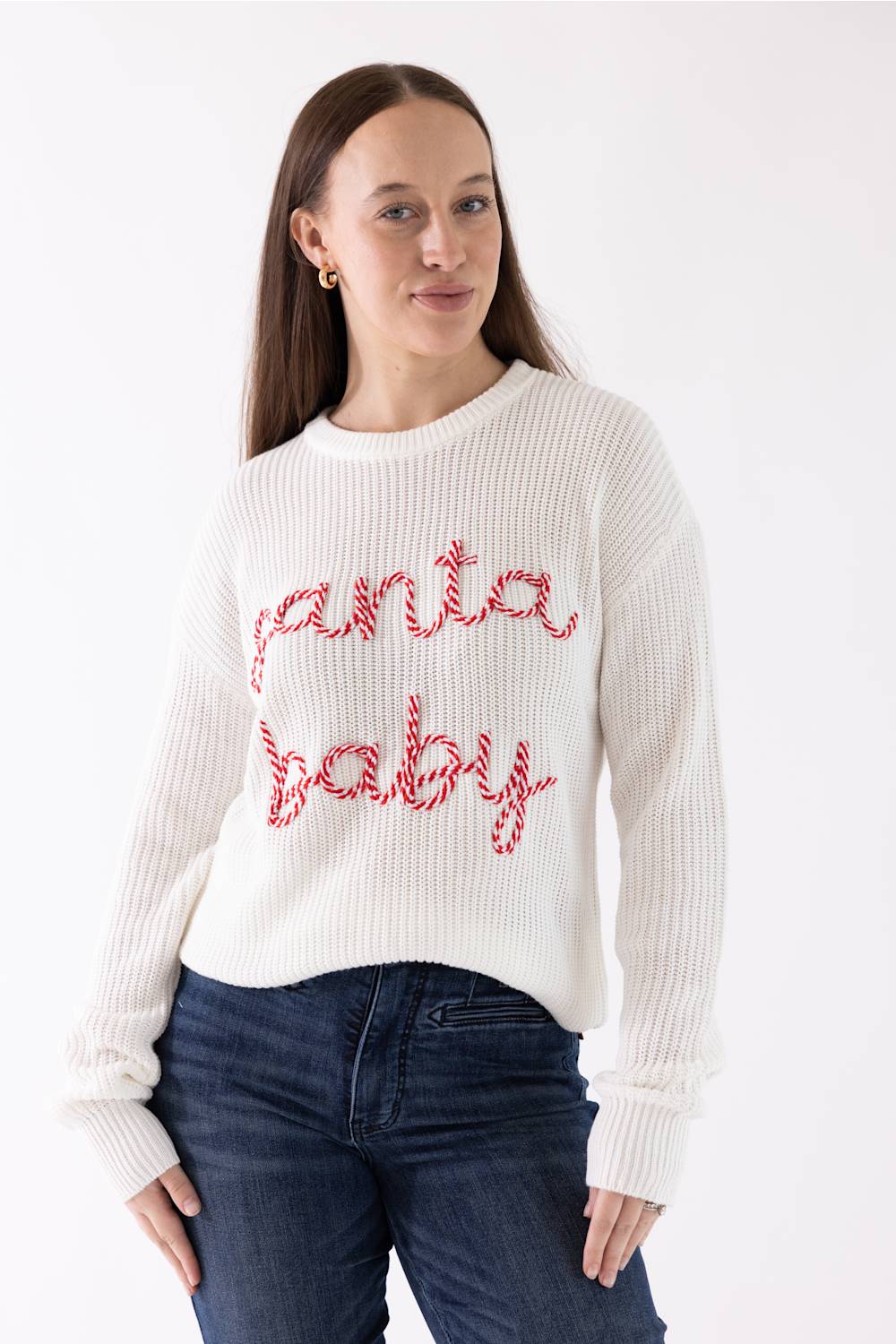 A woman wearing a white ribbed knit sweater with 'Santa Baby' written across the front in red cursive lettering, paired with dark blue jeans, against a plain white background.