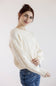 A woman wearing a cream-colored cable-knit sweater and blue jeans, posing against a plain light background.