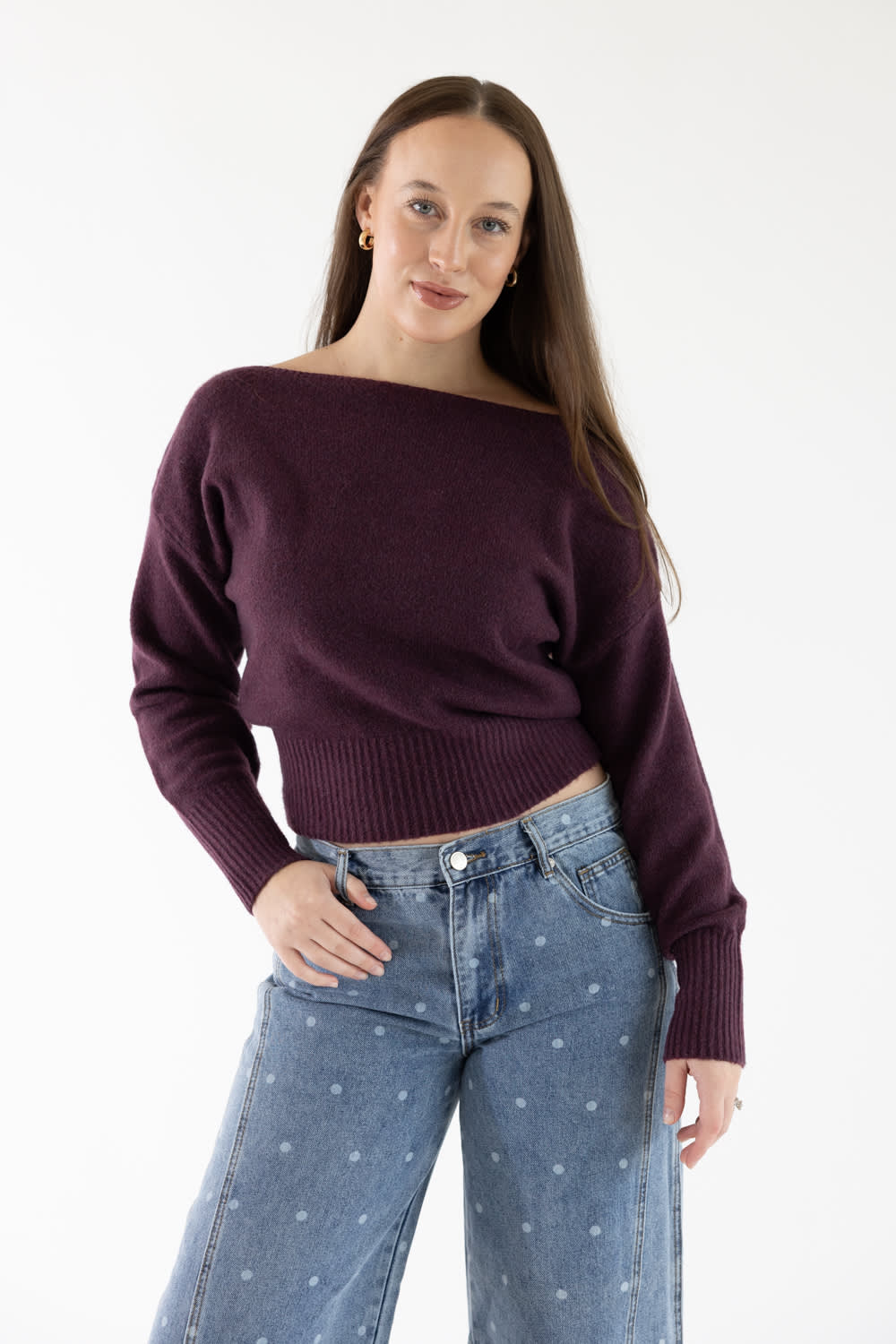 A woman modeling a dark plum purple off-the-shoulder knit sweater paired with blue jeans with a small white star pattern, against a plain white studio background.