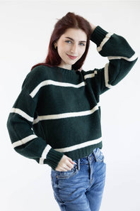 A woman wearing a dark green knit sweater with horizontal cream stripes and blue jeans, posing against a plain white background.