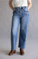 A person wearing light-to-medium wash blue jeans with a straight-leg or wide-leg fit, paired with a white top and brown ankle boots, standing against a plain light gray studio background.