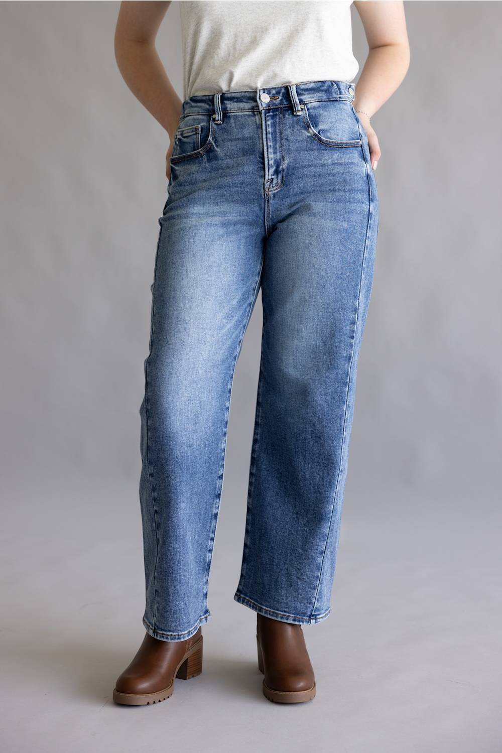 A person wearing light-to-medium wash blue jeans with a straight-leg or wide-leg fit, paired with a white top and brown ankle boots, standing against a plain light gray studio background.