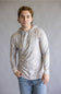 A young man wearing a light gray and white patterned long-sleeve hoodie with blue jeans, standing against a neutral gray studio background.