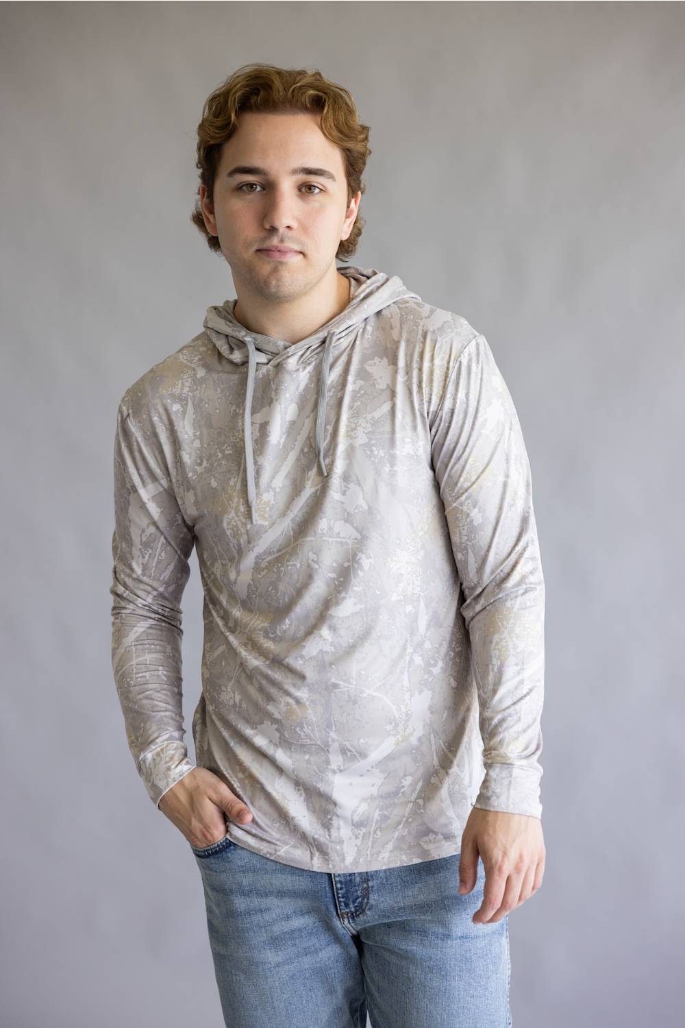 A young man wearing a light gray and white patterned long-sleeve hoodie with blue jeans, standing against a neutral gray studio background.