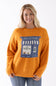 A smiling blonde woman wearing a orange knit sweater with a blue and white pie shop graphic on the front, paired with blue jeans, against a plain white background.
