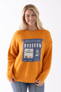 A smiling blonde woman wearing a orange knit sweater with a blue and white pie shop graphic on the front, paired with blue jeans, against a plain white background.