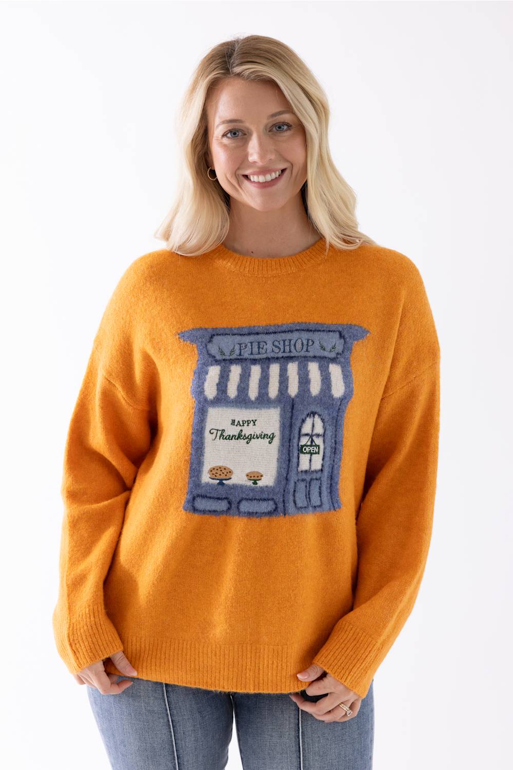 A smiling blonde woman wearing a orange knit sweater with a blue and white pie shop graphic on the front, paired with blue jeans, against a plain white background.