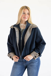 A blonde woman wearing a navy blue jacket with beige and black patterned lining and cuffs, paired with blue jeans, against a plain white background.